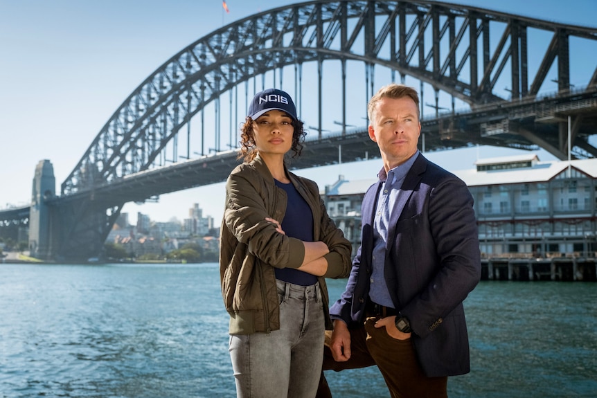 Two actors standing side by side in front of the Sydney Harbour Bridge with one wearing a cap with NCIS written on it