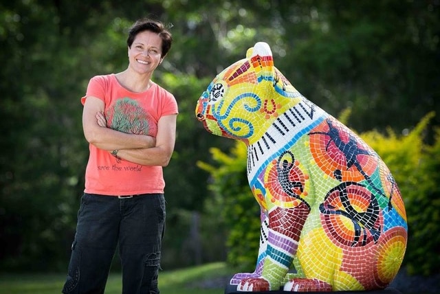 A woman in a t-shirt and jeans stands next to a mosaic koala sculpture, outdoors.