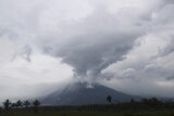 A conical volcano erupts sending thick grey smoke and material into the sky. 