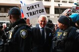 Roger Stone smiles and waves as a protester holds a sign reading Dirty Traitor above his head