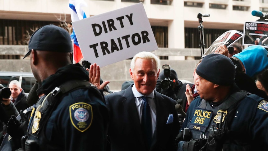 Roger Stone smiles and waves as a protester holds a sign reading Dirty Traitor above his head