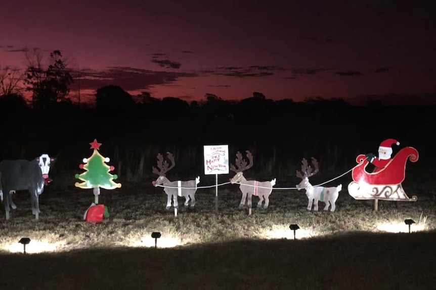 Santa in a sleigh is being pulled by metal reindeer and being watched by a cow at nighttime.
