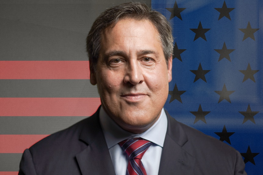 A close up of a man with in a suit & tie smiling at the camera. Around him are the stars and stripes of the US flag.