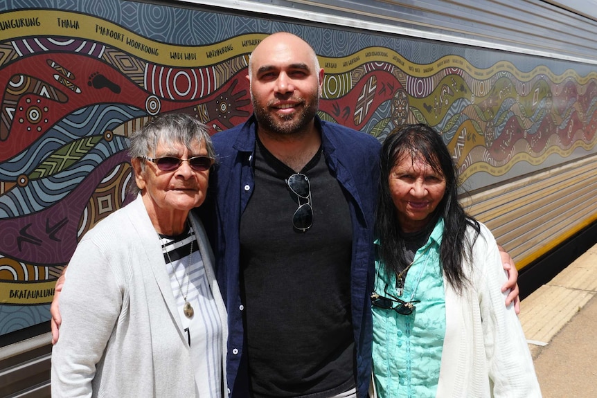 A man stands in front of a train painted with Indigenous artwork with his arms around two older women.