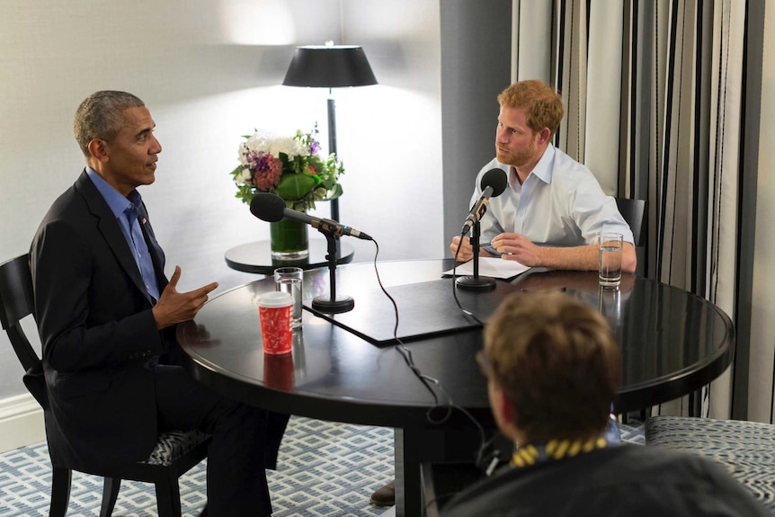 Barack Obama and Prince Harry sit opposite each other at a round table as an interview takes place.