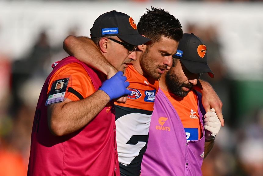 Stephen Coniglio of the Giants is carried off the ground, with a trainer under each arm to help