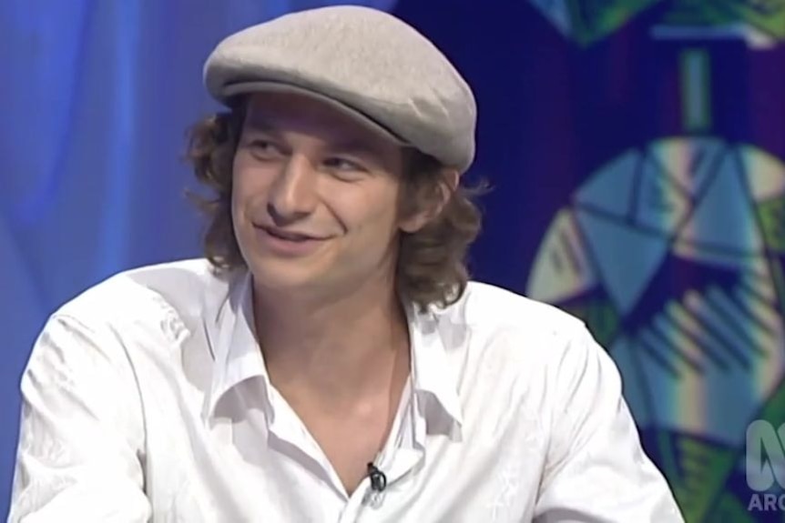 A man in a cap and white shirt speaks in a television studio during a show's filming.