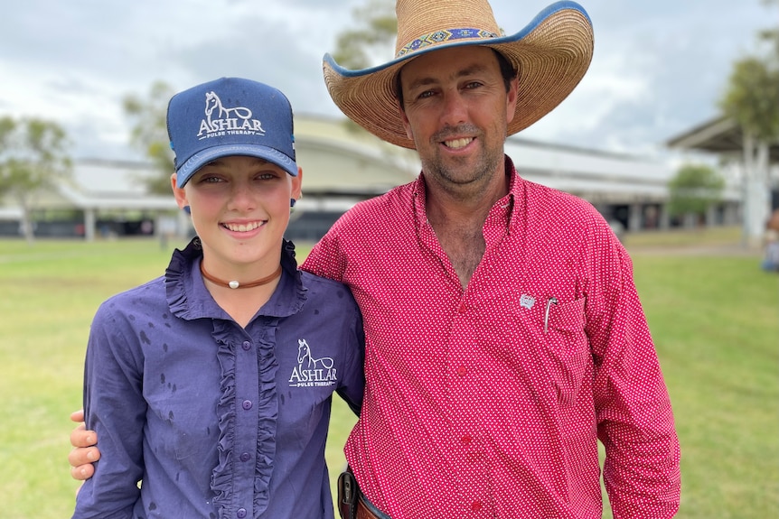 A girl in a purple shirt and cap stands beside a man in a pink shirt.