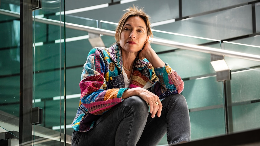 Woman in a multi-coloured and patterned jacket and jeans sits atop some stairs in a modern glass wall space 