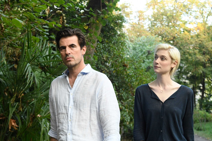 In a garden in the daytime a blonde woman walks behind serious looking tall dark haired man in white cotton button up shirt.