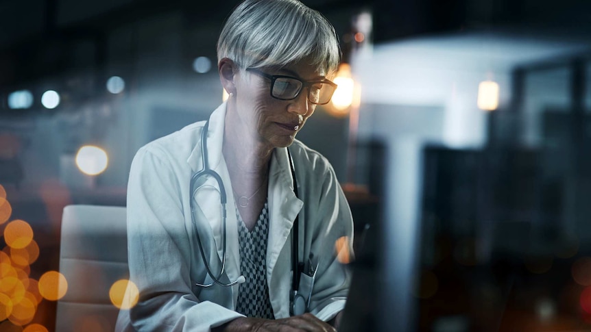 A woman wearing a white coat and stethoscope works on a computer in a darkened room.
