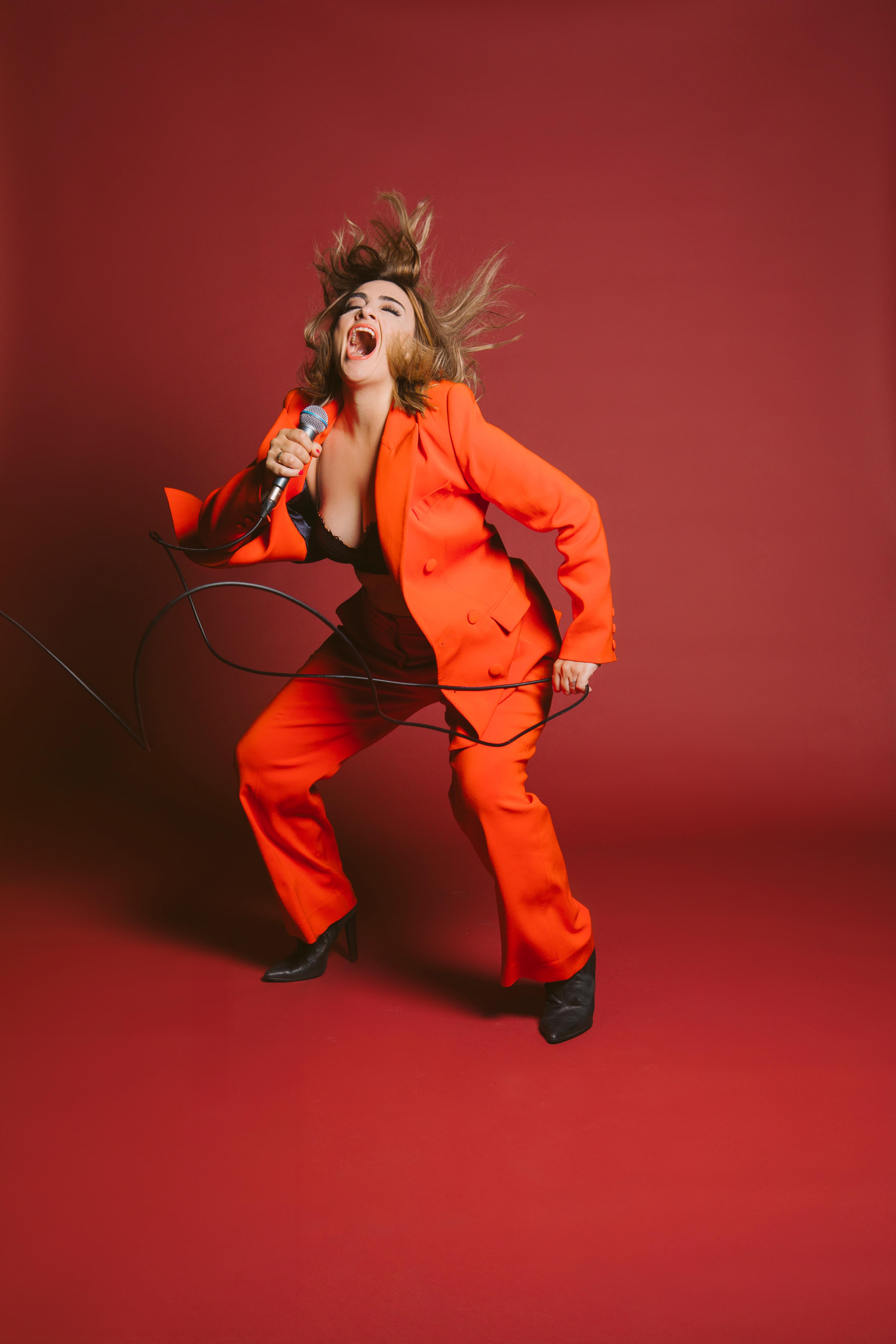 Woman in red jacket and black singlet holds microphone, shaking hair in the air, and with mouth wide open as if singing.