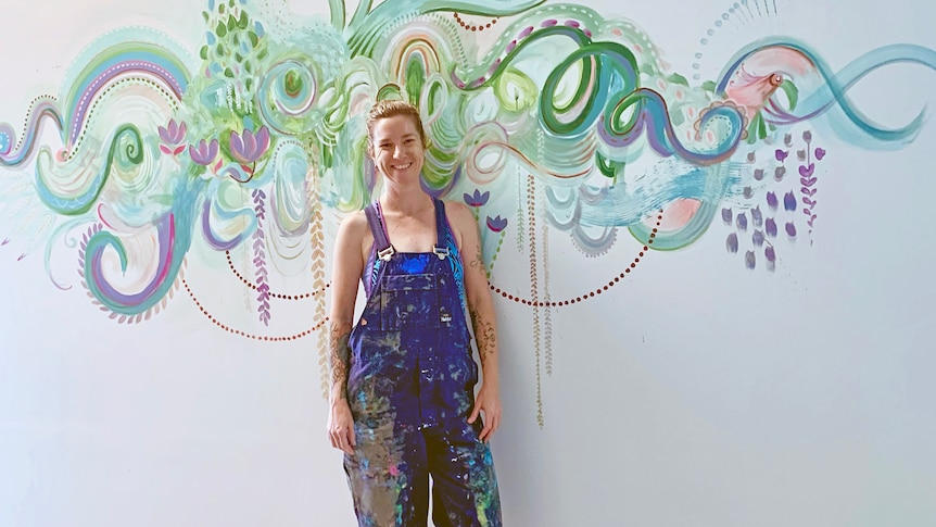 A 42-year-old woman with hair pulled back and wearing paint-splattered blue overalls stands in front of a colourful mural.