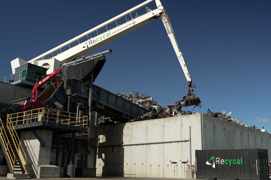 A crane lifts scrap metal onto a large pile with concrete walls around it.