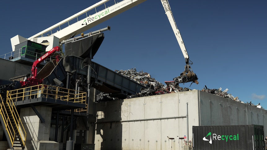 A crane lifts scrap metal onto a large pile with concrete walls around it.