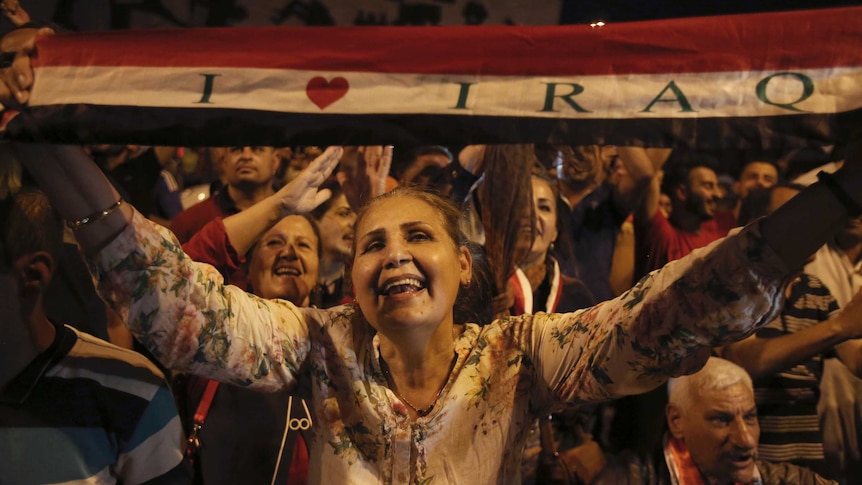 Iraqis celebrate in Tahrir square while holding national flags, smiling. A woman holds a flag that says 'I love Iraq'.