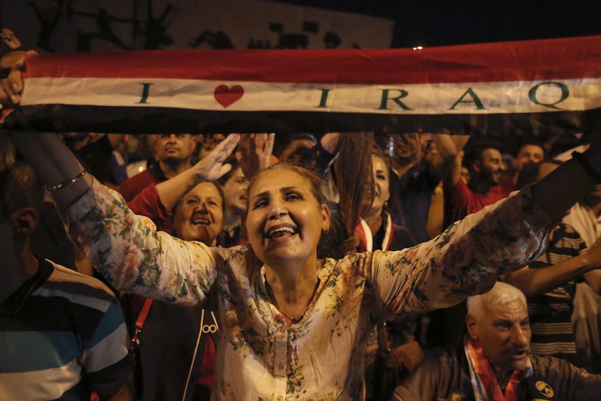 Iraqis celebrate in Tahrir square while holding national flags, smiling. A woman holds a flag that says 'I love Iraq'.