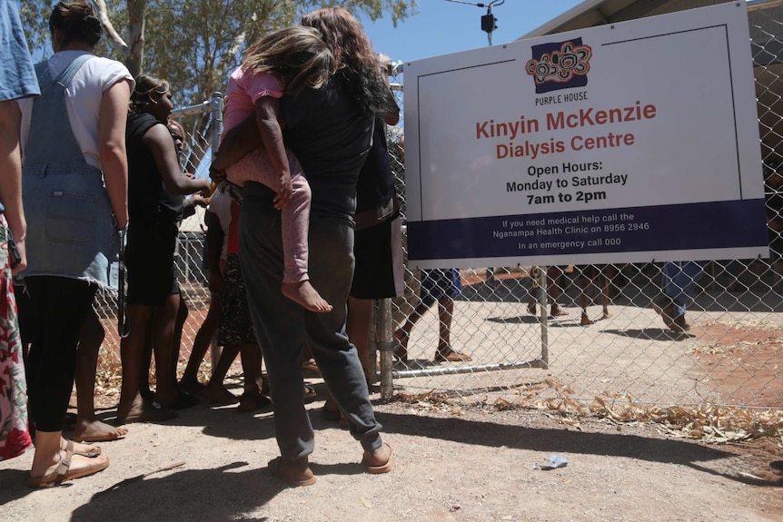 People queue outside a fence which features a sign that reads 'Kinyin McKenzie Dialysis Centre'.
