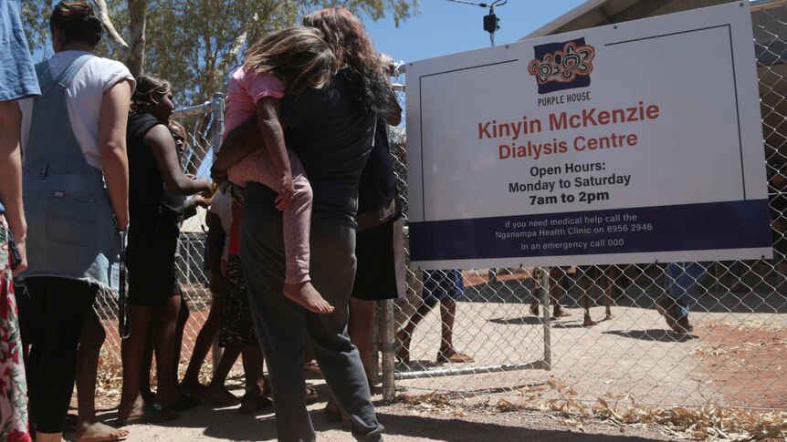 People queue outside a fence which features a sign that reads 'Kinyin McKenzie Dialysis Centre'.