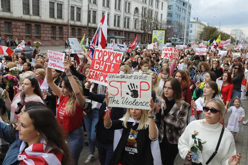 A large group of women march down a main road in daylight while holding signs displaying messages like "proud to be Belarusian".