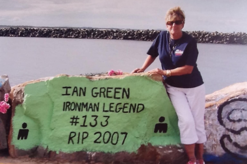 A woman stands next a rock on a breakwall, painted green with words in memory of Ian Green.