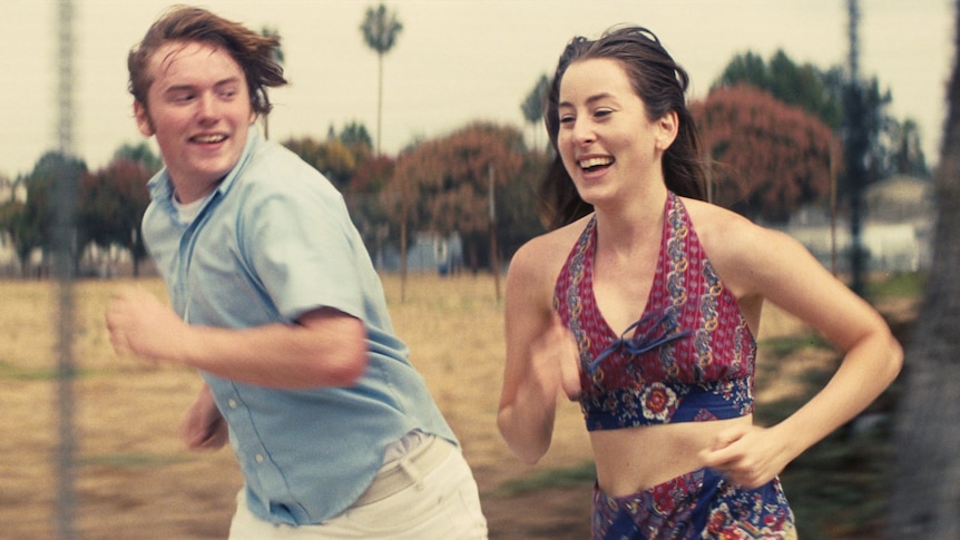 A teenage boy runs excitedly along beside a smiling 20-something brunette woman, each dressed in 70s-style clothing