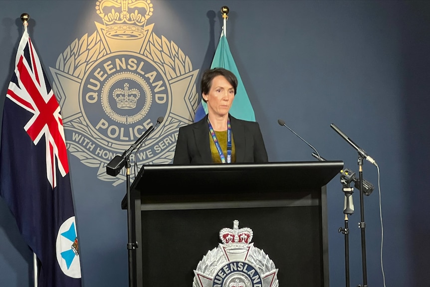 a female police officer wearing a black blazer and a lanyard standing behind a lectern in front of a police badge branded wall