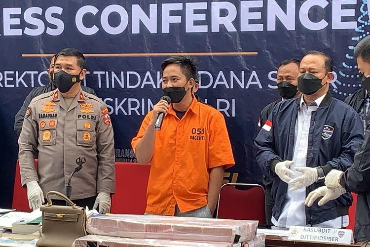 A man wearing an orange prison suit and a face mask stands holding a mic accompanied by two police officers.