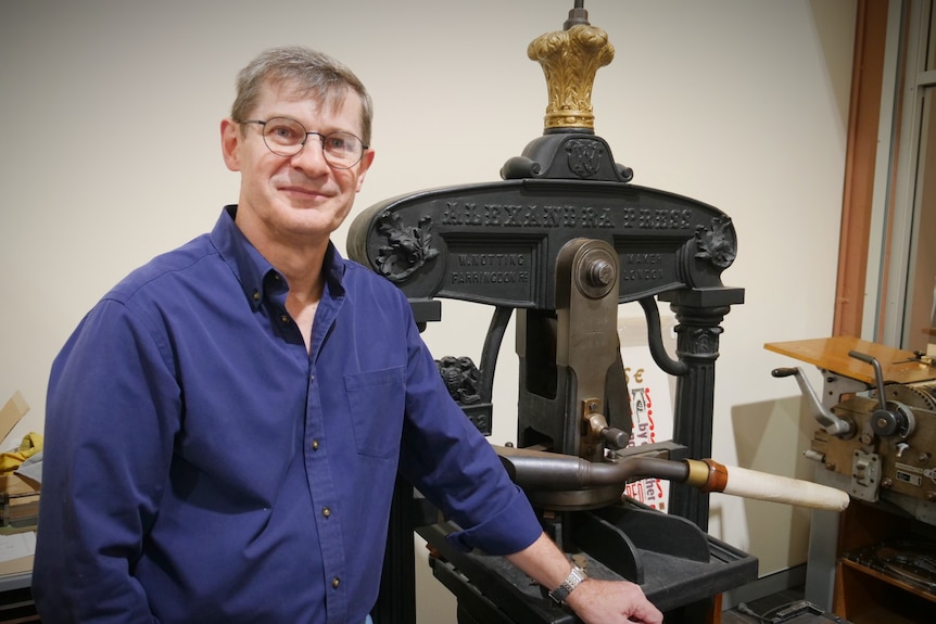 A man in a blue shirt smiles at the camera, he stands next to a vintage printing hand press made of cast iron