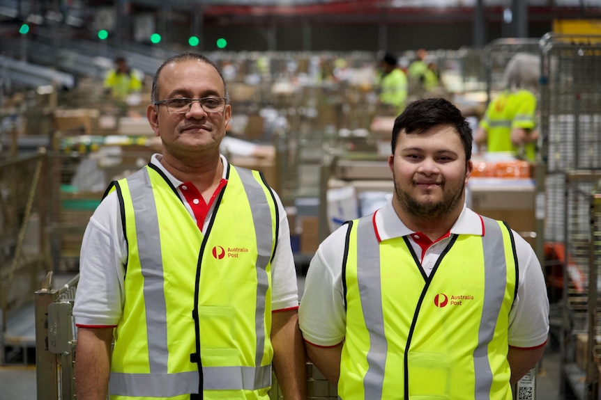 Two men, one older and one young, stand wearing Australia Post high-vis gear.