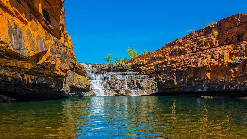 A gorge in the Kimberley with orange rocks around a pool of water, with a waterfall at the end.