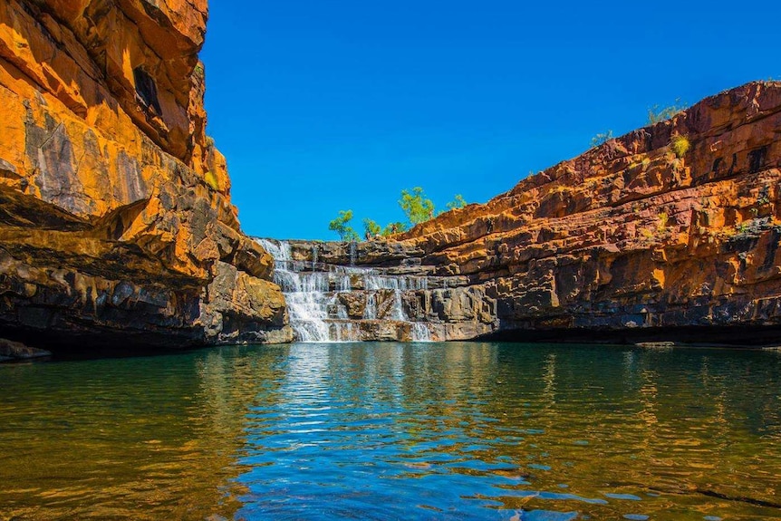 A gorge in the Kimberley with orange rocks around a pool of water, with a waterfall at the end.