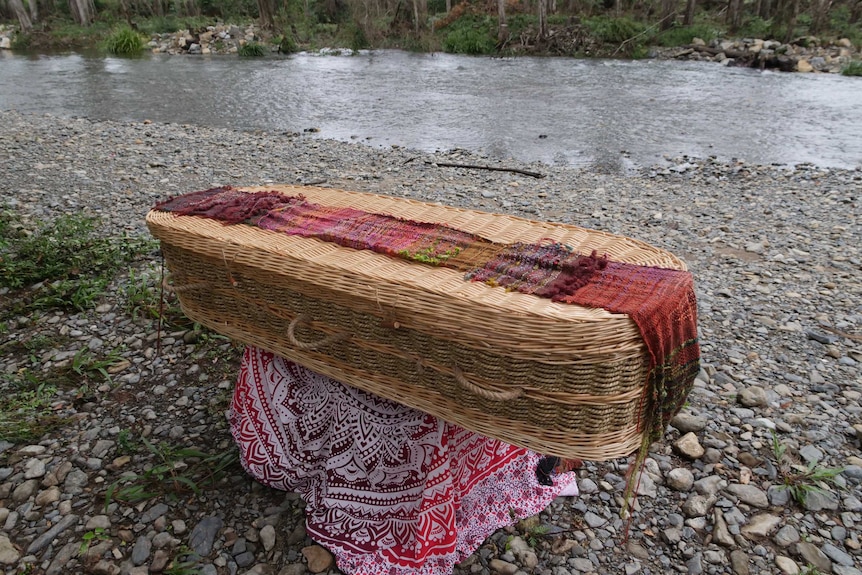 Woven wicker basket coffin with a lumpy hand woven wool run along the top sitting on a stand on the rocks with river behind.
