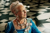 Queen Margrethe wears a crown and ornate necklace with sash over her blue gown while seated for portrait