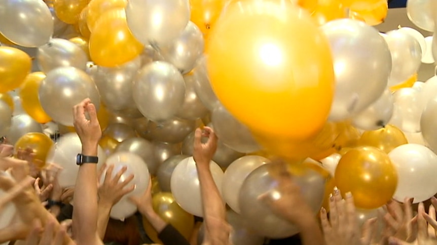 A bunch of yellow balloons with people reaching up for them.