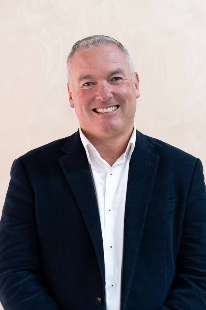 A man wearing a black suit jacket and white shirt smiles for camera.