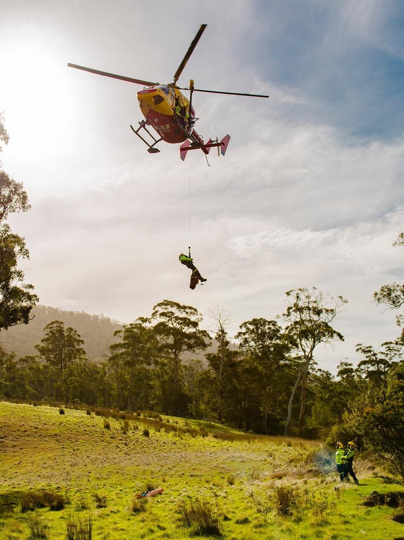 Westpac Rescue Helicopter winching crew member over Tasmanian bush clearing.
