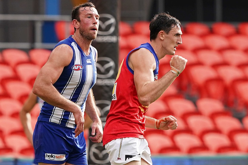 A Brisbane Lions AFL player pumps his right fist as he celebrates a goal with a North Melbourne opponent in the background.