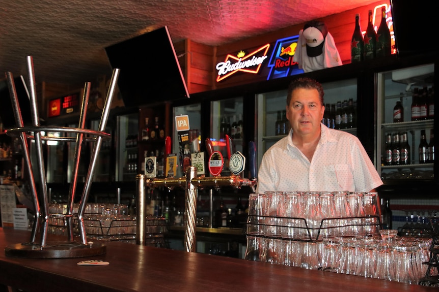 A man stands at a bar with racks of glasses and stools in front of him.