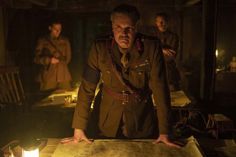 A man in world war one General uniform stands with map and lantern on desk in dim underground room in front of two soliders.