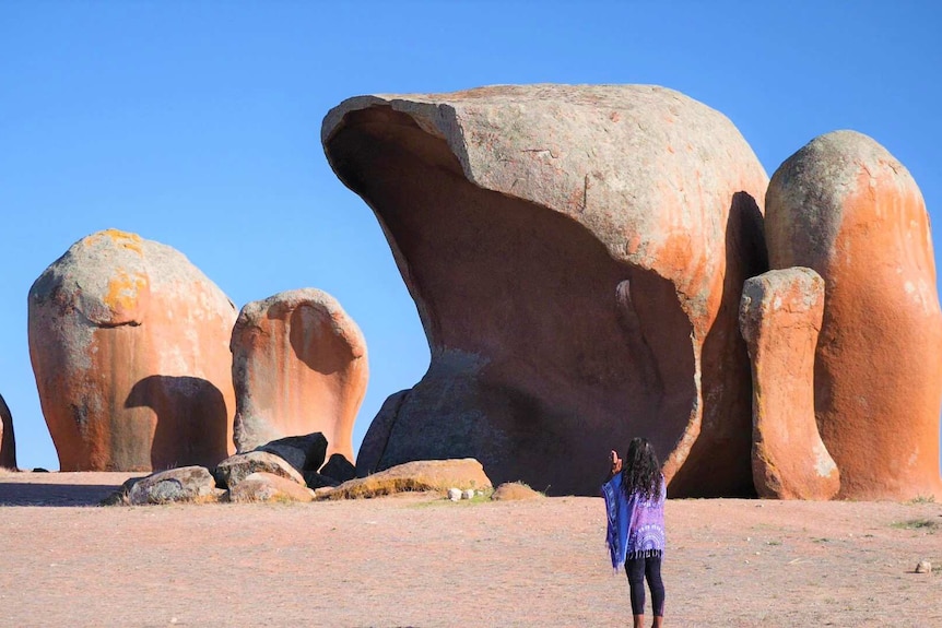 Large boulder rock formation with woman in the foreground hands up in prayer, eagle shadow on rock