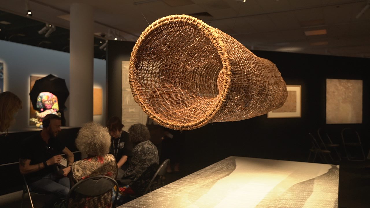A large cylindrical thatched artwork hangs from the ceiling of an art gallery. 