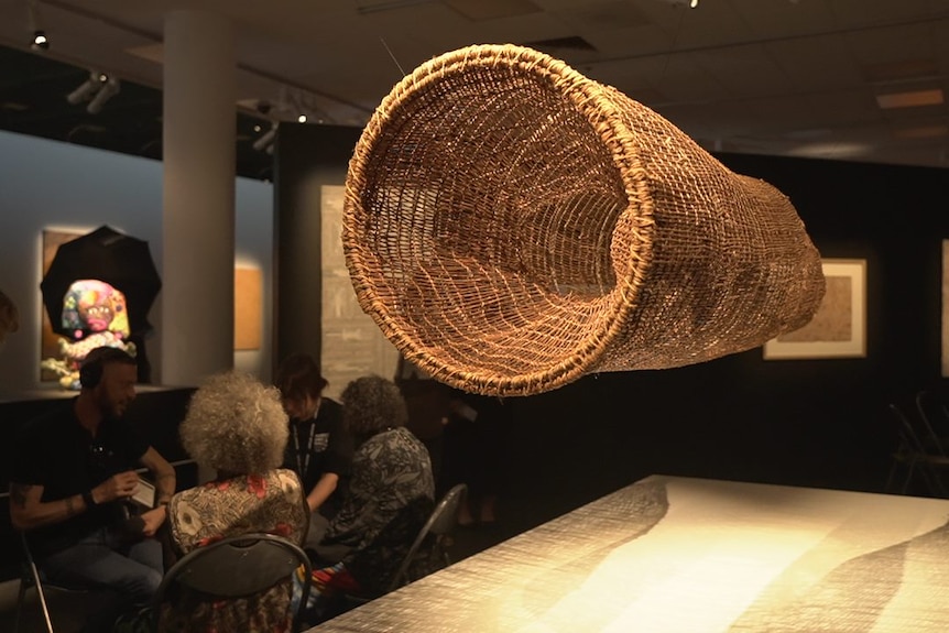 A large cylindrical thatched artwork hangs from the ceiling of an art gallery. 