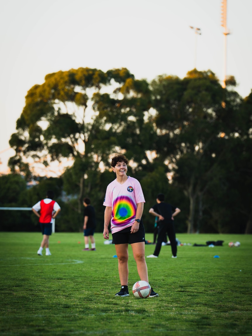 A person wearing a pink shirt with rainbow circles stands on a football field and smiles, with a football at their feet.