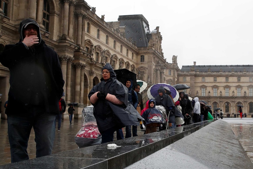 A line of people with umbrellas and raincoats wait outside an old building.