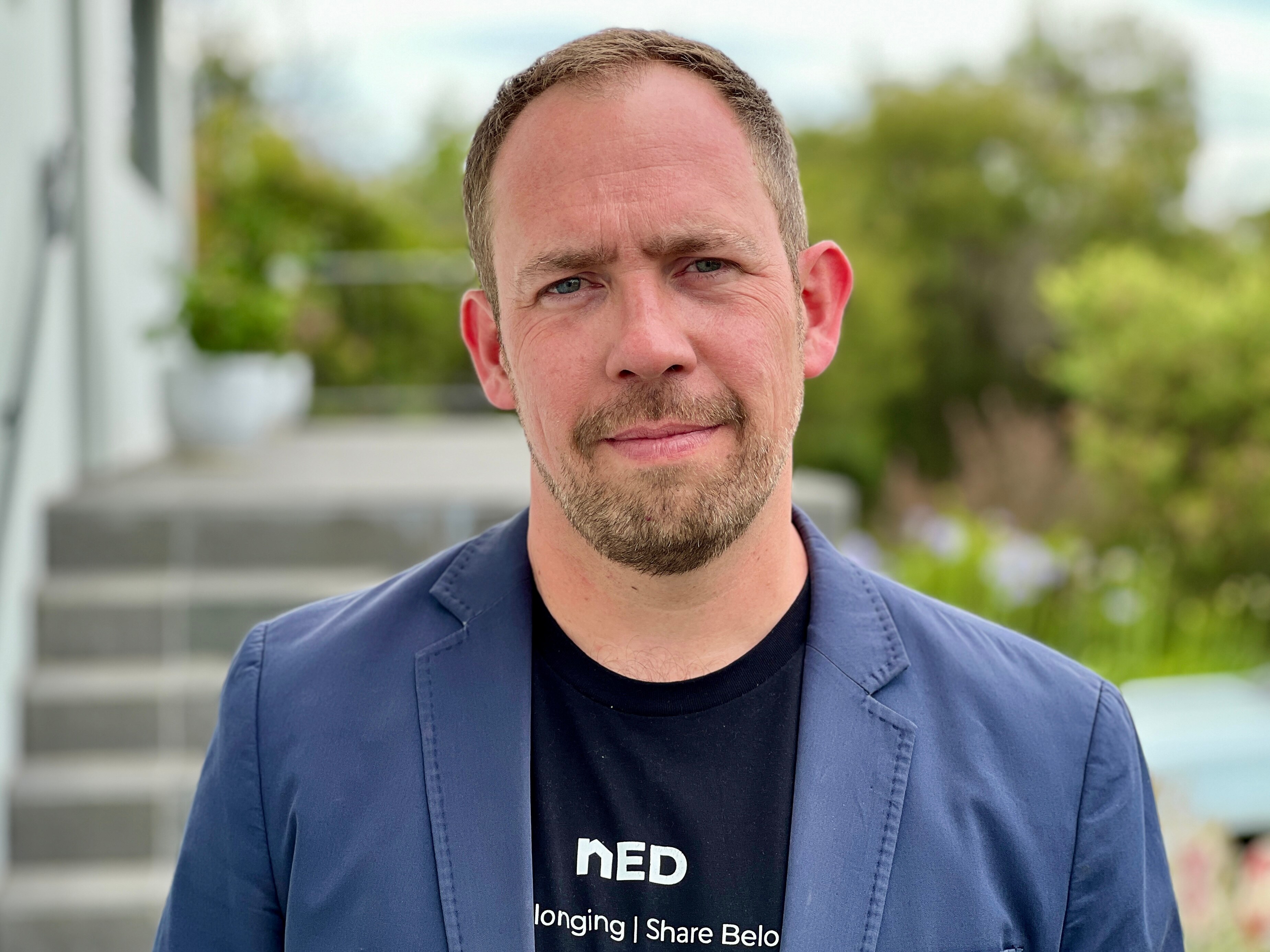 Un hombre blanco con vello facial que vestía una camiseta negra y una chaqueta azul marino.