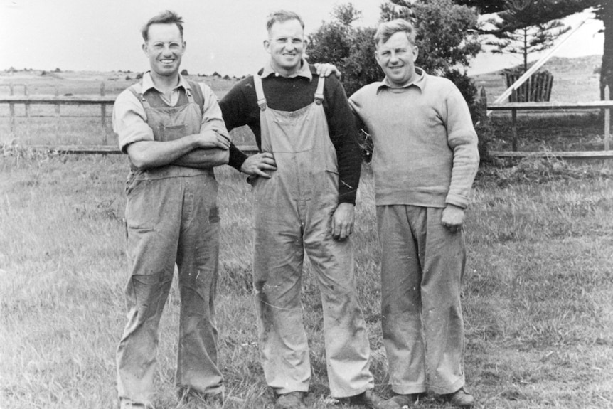 Three men from the 1950s wearing overalls standin in a grassed paddock with a fence behind them, man on left crossing his arms.