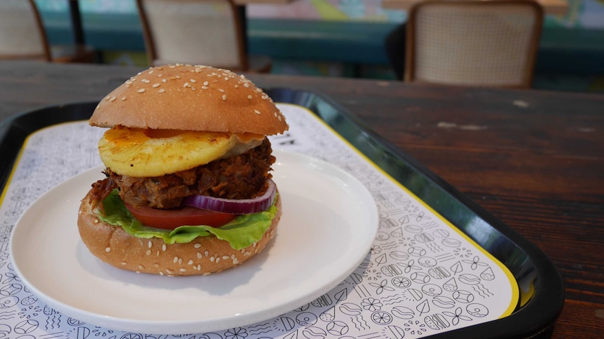 Picture of a burger bun and filling on a plate on a cafe table, with diner out of focus in the background.