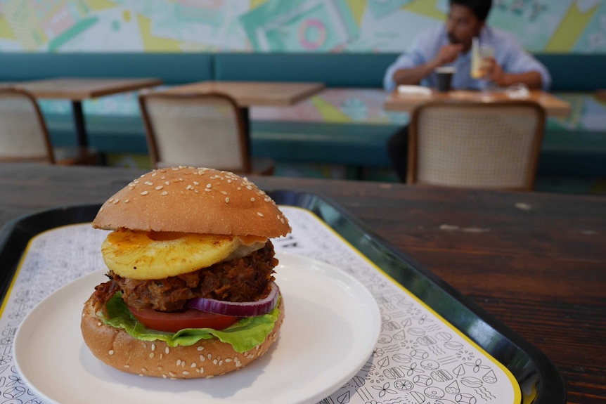 Picture of a burger bun and filling on a plate on a cafe table, with diner out of focus in the background.