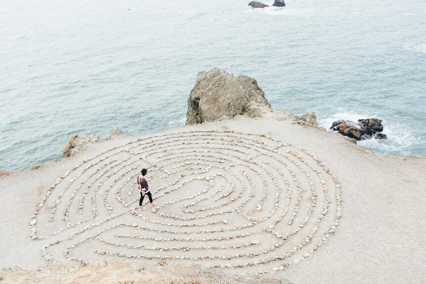 A woman walks through a circular maze made of rocks, that is resting on an ocean-side cliff.